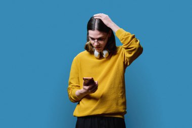 Üzgün adam akıllı telefona bakıyor, mavi stüdyo arka planında kafasını eliyle tutuyor. Duygular, ifade, iletişim, yaşam tarzı, gençlerin kavramı