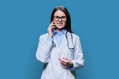 Genç bayan doktor cep telefonuyla konuşuyor, stüdyonun mavi arka planında. Dost canlısı bir doktor hastayla konuşuyor. Tıbbi personel mesleği, teknolojiler mobil uygulamalar, sağlık hizmetleri tıbbı