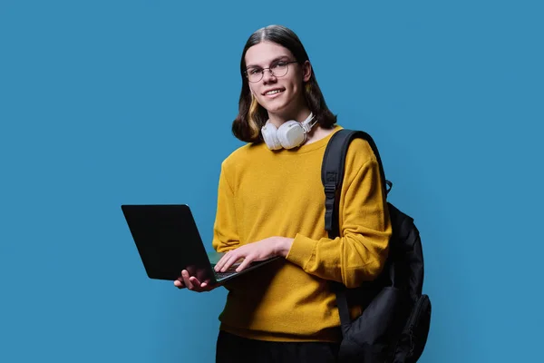Portrait of young male college student using laptop backpack on blue studio background. Handsome trendy guy in glasses headphones looking at camera. Youth, education, high school college university
