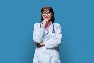 Mavi stüdyo arka planında beyaz önlüklü orta yaşlı kadın doktor portresi. Olumlu gülümseyen olgun tıbbi kadın kameraya bakıyor. Tıbbi personel, meslek, sağlık hizmetleri, bilim hekimliği konsepti