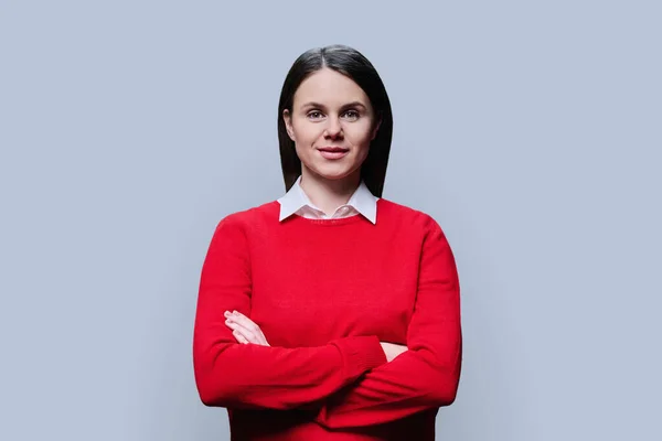 Young smiling woman with crossed arms looking at camera on grey studio background. Beautiful confident successful female in red. Young people 30 years old, business work, lifestyle concept