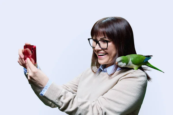 快乐的主人 肩膀上挂着绿和尚鹦鹉 在智能手机上自拍 背景为白色 中年妇女与宠物异国情调鸟类贵格会鹦鹉 关心友情的爱的概念 — 图库照片