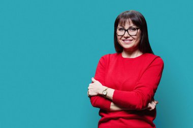 Kırmızı kazaklı gözlüklü, gülümseyen orta yaşlı esmer kadının portresi. Mavi arka planda, fotokopi alanında kameraya bakıyor. Olgun orta yaş, profesyonellik, hizmet, sağlık güzellik uzmanı