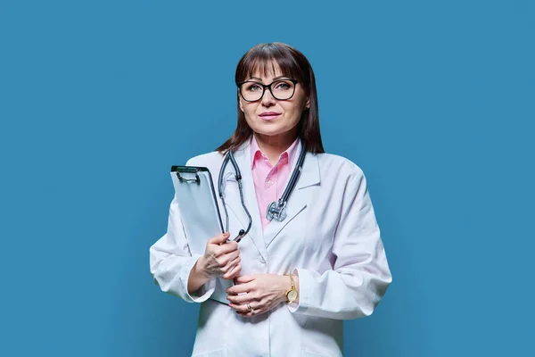 具有蓝色工作室背景的严肃女医生的肖像 中年自信的女人穿着白色外套 带着剪贴板 看着相机 医疗保健 医疗保健 治疗概念 — 图库照片