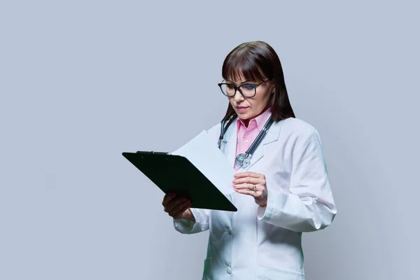 Female doctor with clipboard on gray studio background. Middle aged serious female medic in white coat with stethoscope reading diagnosis on clipboard. Medicine, healthcare, health care, treatment