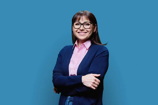 Positive middle aged woman posing over blue studio background. Mature smiling female in glasses with crossed arms looking at camera. Age, profession, job, work, service, business people concept