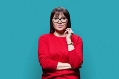 Gülümseyen orta yaşlı esmer kadın portresi kırmızı kazaklı gözlüklü, mavi arka planda kameraya bakıyor. Olgun orta yaş, profesyonellik, hizmet, sağlık güzellik uzmanı