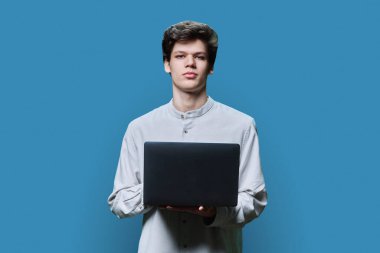 Mavi stüdyo arka planında dizüstü bilgisayar kullanan ciddi bir üniversiteli gencin portresi. 18-20 yaşlarında yakışıklı bir erkek, kameraya bakıyor. Çevrimiçi internet teknolojileri, uzaktan öğrenim