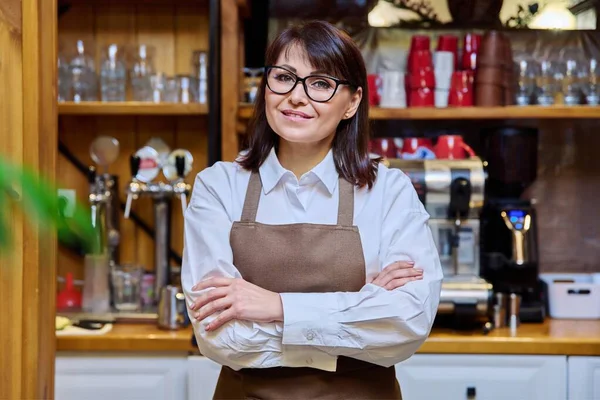 Portrait of middle aged woman bistro restaurant manager owner in an apron, confident smiling looking at camera with crossed arms. Small business, service, work, staff concept