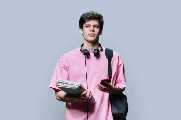 Young guy college student with smartphone backpack, headphones, textbooks, looking at camera on grey studio background. Handsome teenage guy in pink t-shirt, with curly hairstyle. Education technology
