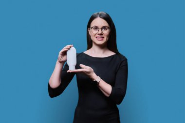Young woman holding white clean tube of body face cream, on blue studio background. Advertising sale of cosmetics for care, treatment, dermatology, cosmetology, beauty industry concept