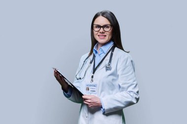 Young friendly woman doctor with clipboard on light gray studio background. Confident female in white coat with glasses looking at camera. Medical staff occupation health care science medicine concept