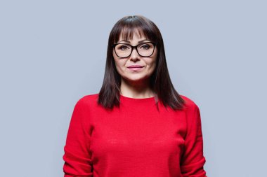 Kırmızı kazaklı gözlüklü, gri arka planda kameraya bakan orta yaşlı esmer kadının portresi. Olgun orta yaş, profesyonellik, hizmet, sağlık güzellik uzmanı