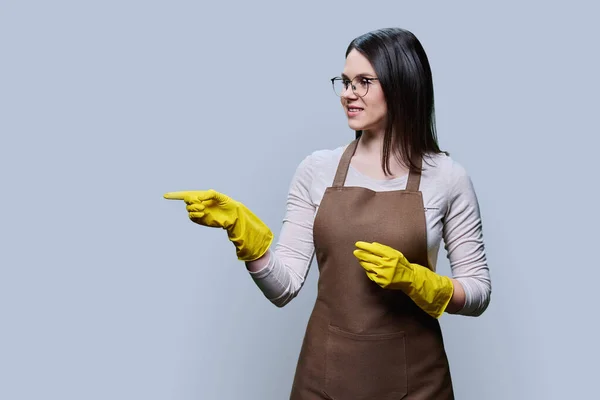 Young woman in rubber glove apron pointing presenting with her hand at empty copy space for text, on grey background. Positive confident female cleaning service worker, smiling housewife