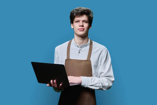Młody Mężczyzna Fartuchu Laptopem Patrzący Kamerę Tle Niebieskiego Studia Praca — Zdjęcie stockowe
