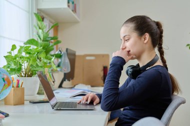 Evde çalışan genç kız, pencerenin yanındaki masada oturan dizüstü bilgisayarla çalışıyor. Genç bir bayan ekrana bakıyor, uzaktan çalışıyor, online bir ders dinliyor, ödevlerini yapıyor.