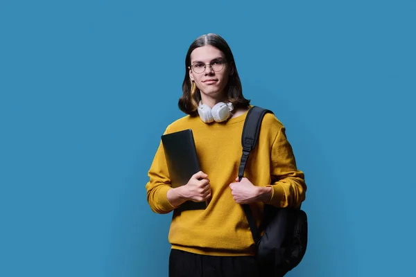 Portrait of young male college student with laptop backpack on blue studio background. Handsome trendy guy in glasses headphones looking at camera. Youth, education, high school college university