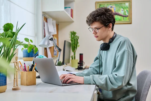年轻的男大学生坐在家里的办公桌前 用笔记本电脑 一个19岁的帅哥 戴着眼镜在键盘上打字 利用互联网在线技术进行学习 — 图库照片