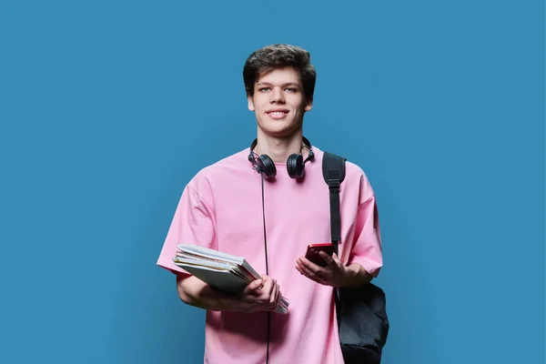 Young guy college student with smartphone backpack, headphones, textbooks, looking at camera on blue studio background. Handsome teenage guy in pink t-shirt, with curly hairstyle. Education technology