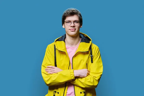 Handsome guy posing in outwear, on blue studio background. Trendy young male in knitted hat yellow jacket, looking at camera with arms crossed. Seasonal spring autumn winter clothing, fashion, youth