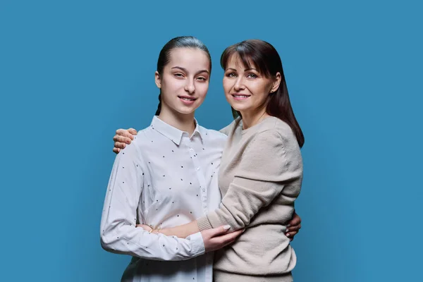 Portrait of middle aged mother and teenage daughter embracing together on blue studio background. Smiling happy mom and girl looking at camera. Family, lifestyle, relationship, mothers day concept