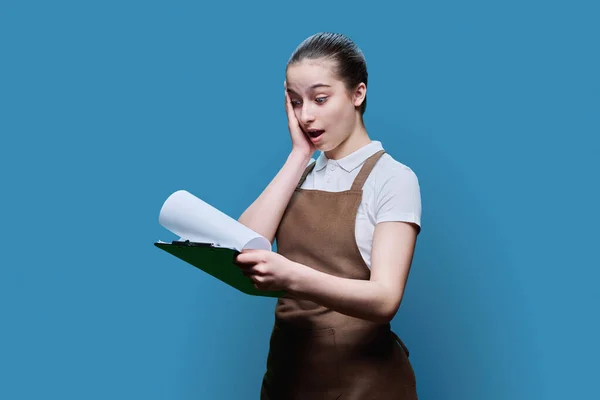 穿着围裙的年轻女服务员看到蓝色工作室背景的剪贴板时 大吃一惊 服务部门的工作 学生就业 咖啡店的工作人员 — 图库照片