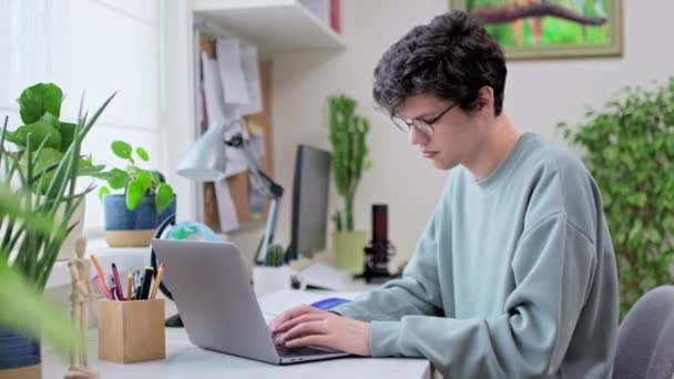 年轻的男大学生坐在家里的办公桌前 用笔记本电脑 一个19岁的帅哥 戴着眼镜在键盘上打字 利用互联网在线技术进行学习 — 图库视频影像