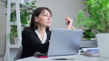 Çok çalışan orta yaşlı bir kadın iş yerinde oturmuş, camdan dışarı bakarken gözlüklerini çıkarıyor. Stres, yorgunluk ve üzüntü yaşayan olgun bir kadın ofiste oturuyor.