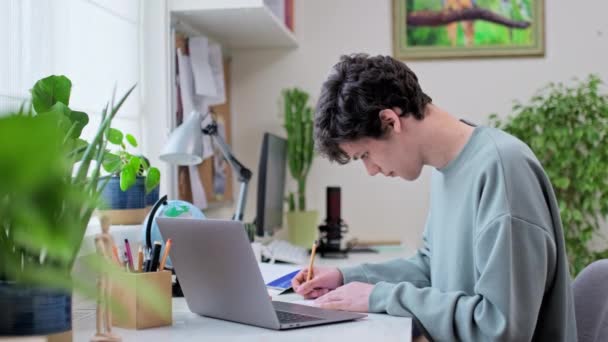年轻的男大学生坐在家里的课桌前 用笔记本电脑学习 英俊的男人19岁看着相机 利用互联网在线技术进行学习 — 图库视频影像