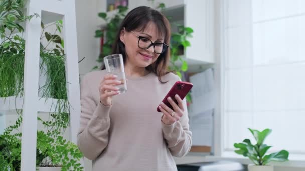 Kobieta Średnim Wieku Szklanką Wody Pomocą Smartfona Wnętrzu Domu Wypoczynek — Wideo stockowe