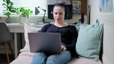 Kulaklıklı genç bir kız dizüstü bilgisayara bakıyor, evdeki kanepede uzanıyor, rahatlama ve iletişim kurmayı öğreniyor. Ciddi özenli genç bayan video izliyor, internet teknolojisi konsepti