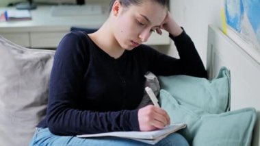 Genç kız evde ders çalışıyor, ders kitabı okuyor, deftere yazıyor, evdeki kanepede oturuyor. Ergenlik, lise öğrencisi, eğitim öğrenme konsepti