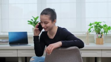 Genç kız telefonda konuşuyor, evdeki sandalyede oturuyor. Genç bayan iletişim eğlence çalışması için telefon kullanıyor.