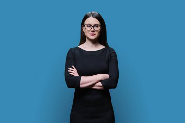Gözlüklü, siyah elbiseli, mavi stüdyo arka planında poz veren güzel bir kadının portresi. Kollarını kavuşturup kameraya bakan kendinden emin bir kadın. Moda, güzellik, vücut bakımı, satış yönetimi uzmanı.