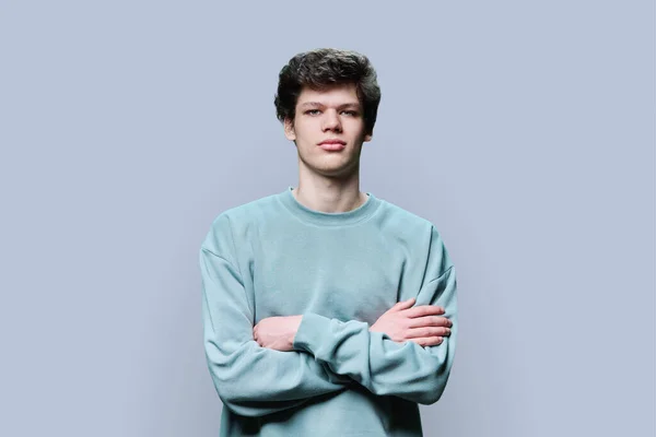 Portrait of serious calm young male 18-20 years old in casual sweatshirt with crossed arms on gray studio background. Handsome confident guy with curly hair looking at camera. Lifestyle, youth concept
