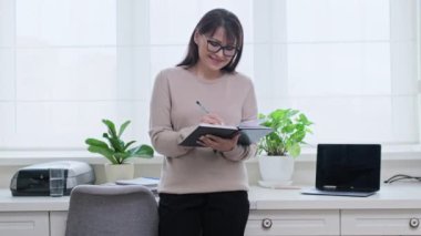 Not defterine not alan orta yaşlı bir kadın, masanın yanında duruyor, ofisin arka planında.