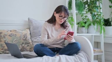 Evdeki koltukta oturan orta yaşlı kadın akıllı telefon ve dizüstü bilgisayar kullanıyor. Uzaktan çalışma, serbest çalışma, eğlence, yaşam tarzı konsepti