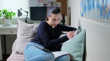 Genç kız evde ders çalışıyor, ders kitabı okuyor, deftere yazıyor, evdeki kanepede oturuyor. Ergenlik, lise öğrencisi, eğitim öğrenme konsepti