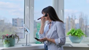 Makyaj yapan orta yaşlı bir kadın aynaya bakarak allık tozu bronzlaştırıcısı sürüyor. Güzellik, kozmetik, cilt bakımı, makyaj, olgun kadın konsepti