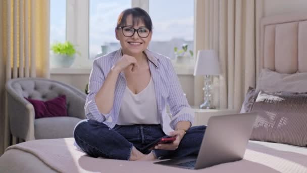 積極的な中年の女性のベッドの上にノートパソコンのスマートフォンを使用して自宅に座って カメラを見てポーズ 歯で笑顔 ライフスタイル レジャー フリーランス 成熟した人々の概念 — ストック動画