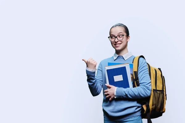 Teen girl high school student pointing finger sideways space for text on white studio background. Smiling female in blue sweater glasses with backpack looking at camera. Education, adolescence