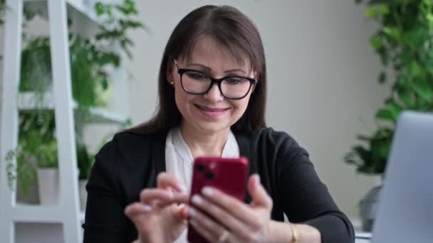 レジャーのためのスマートフォンを使用して職場のホームオフィスで中年の女性 笑顔の女性見る正ビデオを読む良いニュース — ストック動画