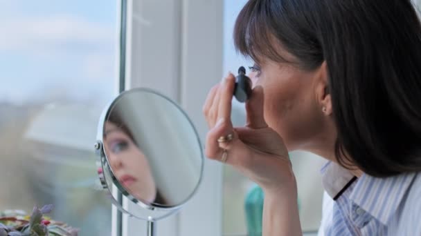 鏡を見てまつ毛にマスカラを適用し メイクを行う中年の女性は 自宅でウィンドウ 化粧品 成熟した女性の概念 — ストック動画