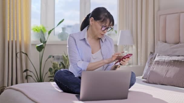 中年妇女拿着智能手机拨号在触摸屏上 看笔记本电脑 在家里的床上沙发上打电话 通信网络生活工作休闲 成熟的人 — 图库视频影像