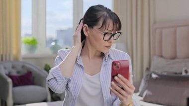 Evdeki koltukta oturan orta yaşlı bir kadın, cep telefonunun ön kamerasında kendine bakıyor, saçını düzeltiyor, kendisine hayran. Yaş, yaşam tarzı, güzellik, olgun insanlar konsepti