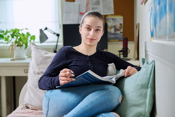 Teenage girl studying at home, reading textbook, writing in notebook, sitting on couch at home. Adolescence, high school student, education learning knowledge concept