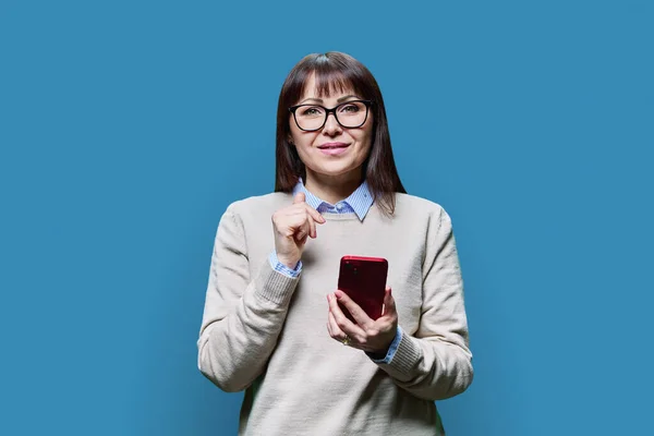 stock image Serious mature woman with smartphone in hands looking at camera on blue studio background. Middle aged female in casual jumper. Work, staff, freelancing, technology lifestyle leisure concept