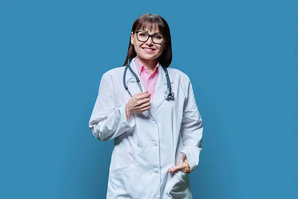 中年妇女医生的画像 身穿白色外套 背景为蓝色 正笑着看着成熟的医学女性镜头 医务人员 科学医学概念 — 图库照片
