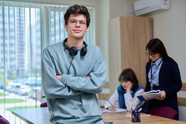 Portrait of college student guy looking at camera inside classroom. Young male 18, 19 years old wearing glasses, with headphones, posing in education center. Education, knowledge, learning, youth