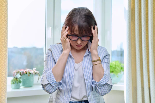 一个成熟的女人头痛 双手抱头 站在窗前 偏头痛 中年健康概念 — 图库照片